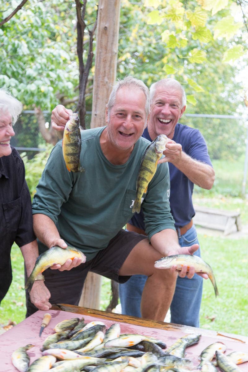 A guest holding up two perch fresh off the pergola picnic table, family looking on